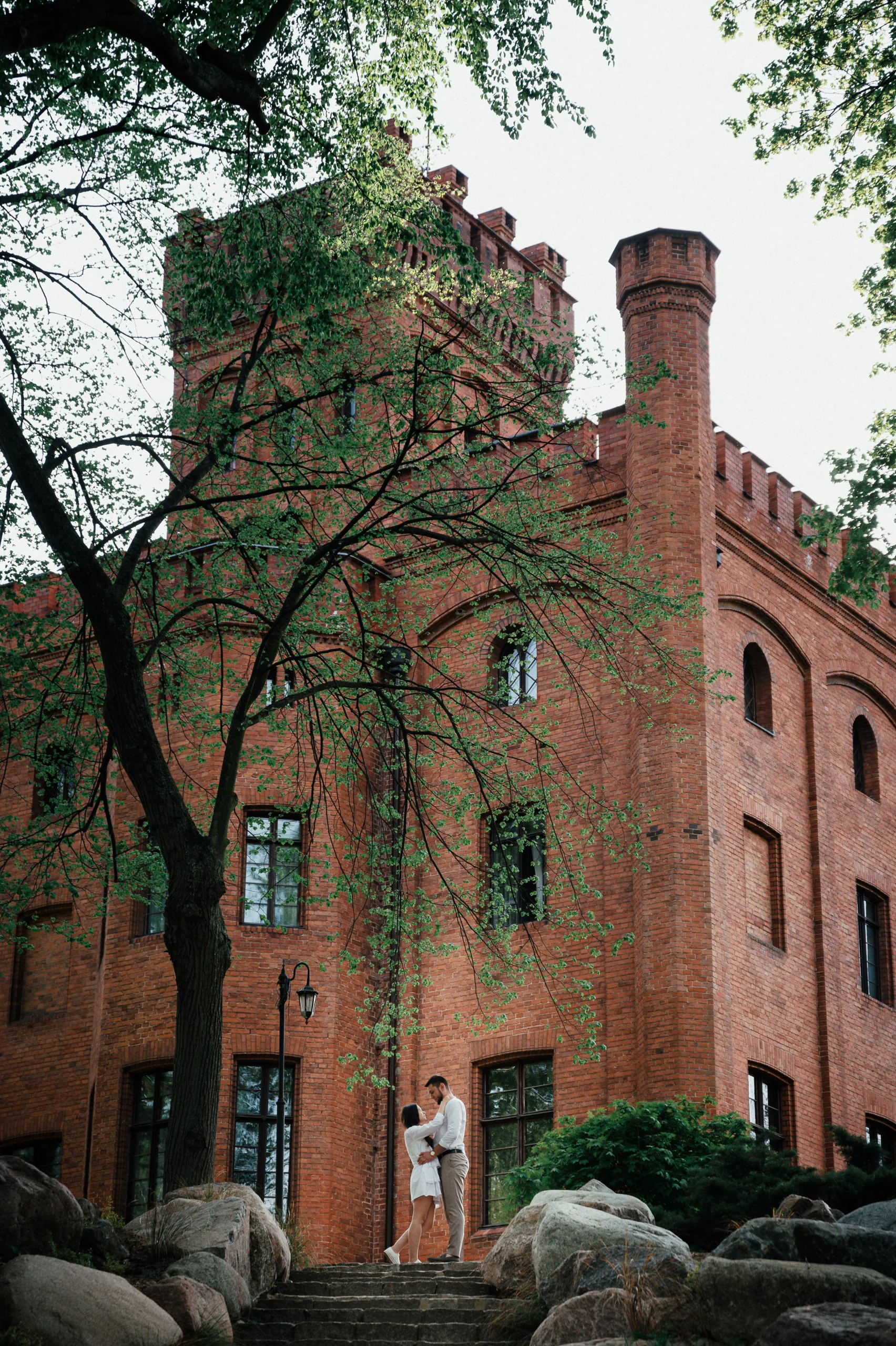 Para młoda przed zabytkowym budynkiem wśród drzew.